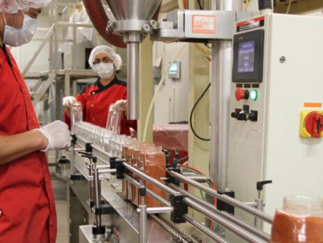 Image of Majestic Spice production line. Two Majestic Spice workers overseeing the automated bottling process of spice jars. Both of them are wearing a red shirt, hairnets and white gloves, placing empty glass jars onto a conveyor belt.The jars appear to be filled with a reddish spice blend. The Majestic Spice logo is displayed in the bottom left corner.