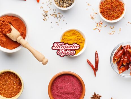 Different spices in different types of rounded containers in a white background. In the center is the logo "Majestic Spice" overlaid on a bright yellow bowl of powder, surrounded by several other bowls and piles of spices including a deep red paprika or chili powder with a wooden scoop, golden turmeric, a mix that appears to be a chili lime blend, and whole red chilies. There are also hints of other seasoning ingredients like garlic cloves, star anise, bay leaves, and nutmeg.