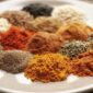 In this image we can see a wooden background and a big white plate with a lot of small bunches of spices like turmeric, chili, some herbs, and other white, black, white, and brown spices. At the bottom right corner, the majestic spice logo is in red.