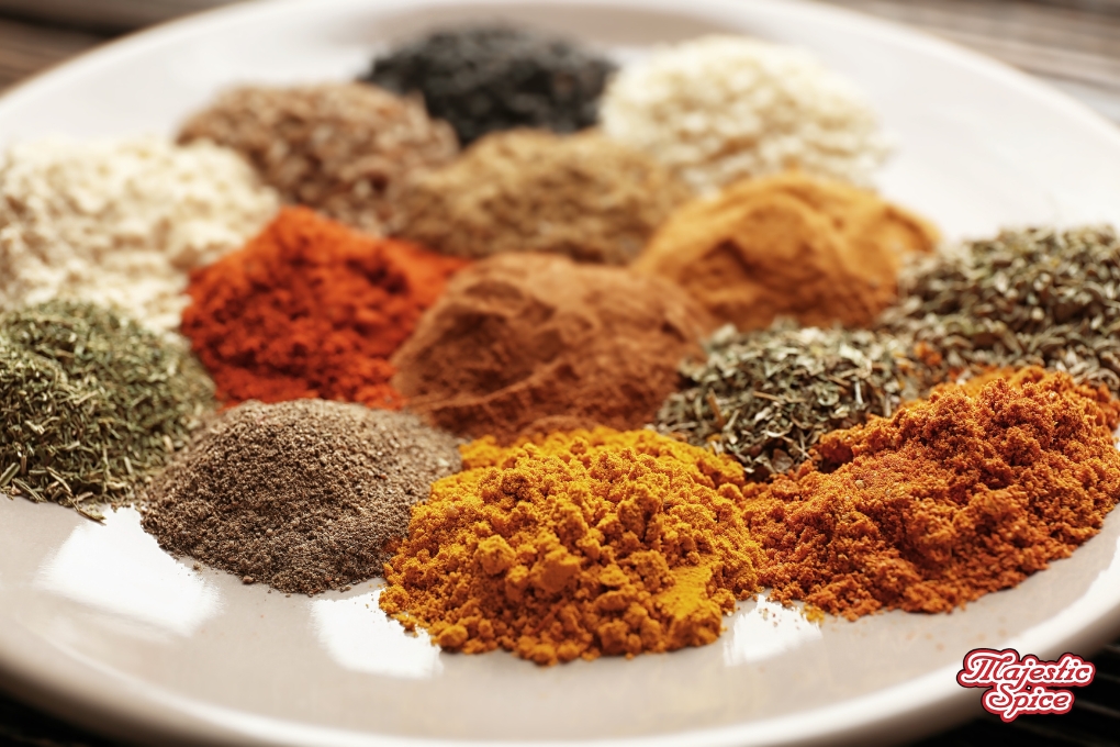 In this image we can see a wooden background and a big white plate with a lot of small bunches of spices like turmeric, chili, some herbs, and other white, black, white, and brown spices. At the bottom right corner, the majestic spice logo is in red.